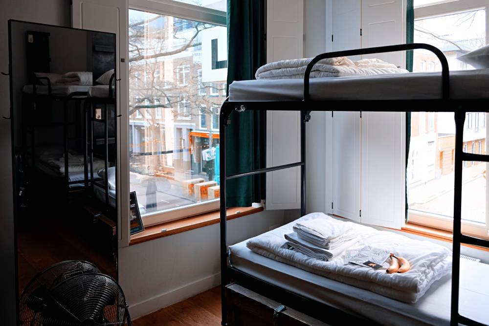 A cozy hostel room featuring a bunk bed with neatly arranged linens, a window with green curtains, and a reflection in a mirror.