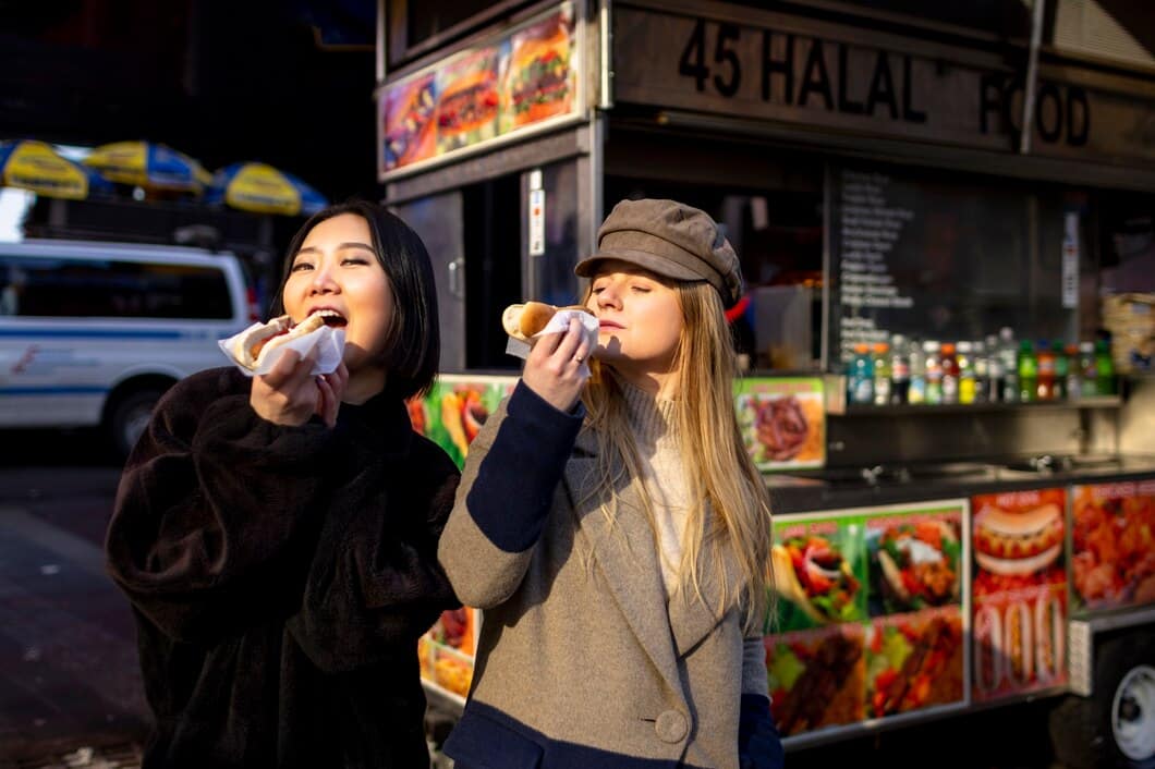 Two women enjoy hot dogs in front of a street food cart with colorful menu photos and a blurred street in the background.