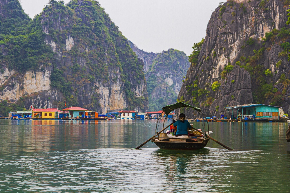 A person rows a wooden boat past colorful floating houses surrounded by tall, rugged cliffs in a serene, misty harbor.