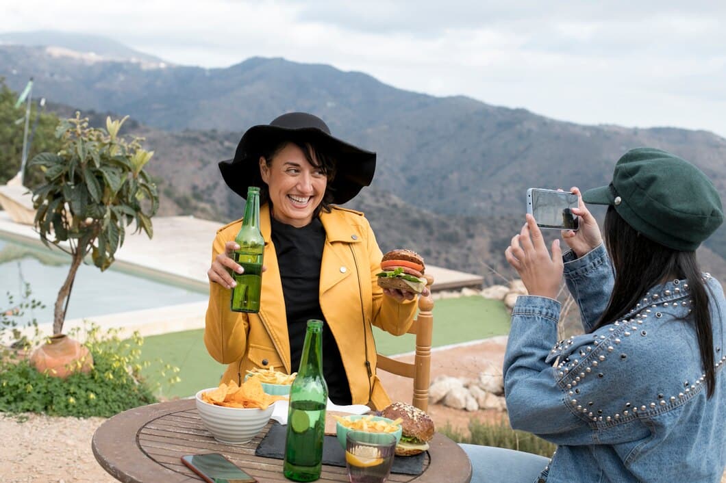 A woman in a yellow jacket holds a burger and drink at a scenic outdoor table, while a friend captures the moment on her phone.
